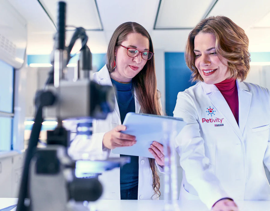 Two female scientists in lab coats discussing notes next to laboratory equipment in a bright research setting.