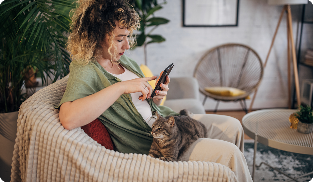 A woman sitting on a cozy couch interacts with her smartphone while a cat rests on her lap in a warmly lit living room.