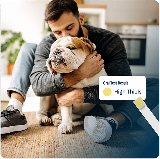 Man sitting on the floor hugging a bulldog, with an oral test result overlay showing 'High Thiols' indicated by a yellow test strip.